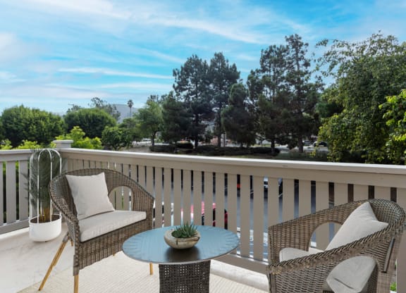 Two-Bedroom Apartments in Lompoc, CA - Oceanwood - Private Balcony with Patio Chairs, Coffee Table, Potted Plant, and View of Parking Lot with Lush Trees in the Background.