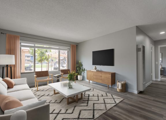 Goleta, CA Apartments Near UC Santa Barbara - Patterson Place - Living Room With Wood-Style Flooring, Area Rug, Couch, Armchairs, Coffee Table, Media Cabinet, Wall-Mounted TV, Standing Lamp, And Large Window.