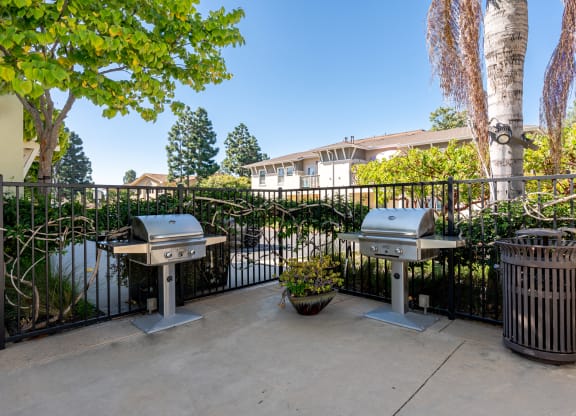 Ventura, CA Apartments Near The Collection Oxnard - Ralston Courtyards - Outdoor BBQ Grills At BBQ Area With Surrounding Steel Railings, Bushes, And Trees.