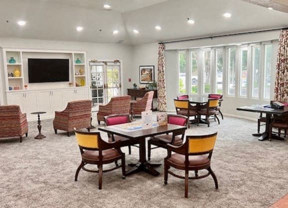 Santa Barbara, CA, Apartments Near Target - Rancho Franciscan - Community Clubhouse With Tables and Chairs, Carpeting, Mounted TV, Bookshelf, and Large Windows