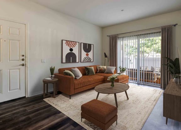 Goleta CA Apartments - Sumida Gardens - Furnished Living Room with Wood-Style Flooring, Area Rug, Burnt Orange Sofa, Ottoman, Wooden Round Coffee Table, Side Table with Plant, Wooden Monitor, Standing Lamp, Framed Art, and Glass Sliding Door with Access to Balcony