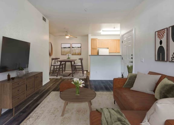 1-Bedroom Apartments in Goleta, CA - Sumida Gardens - Living Room with Wood-Style Flooring, Area Rug, Sectional Sofa, Round Coffee Table, Stool, Media Cabinet, Wall-Mounted TV, and View of Dining Area and Kitchen.