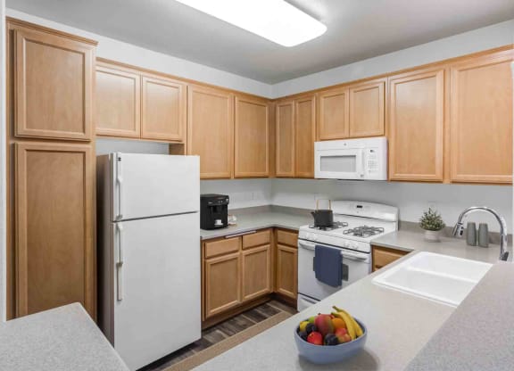 Apartments for Rent in Goleta CA - Sumida Gardens - Kitchen with Wood-Style Floor, Area Rug, White Appliances, Wood Cabinetry, Grey Countertops, Overhead Lighting, and Breakfast Bar Area