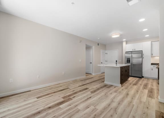 an empty living room with a kitchen and a stainless steel refrigerator