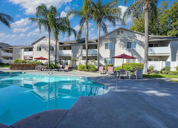 Community swimming pool with large palm trees in the background as well as one of the community apartment buildings. There are tables and chairs around the pool deck at The Legacy Apartments, Antelope
