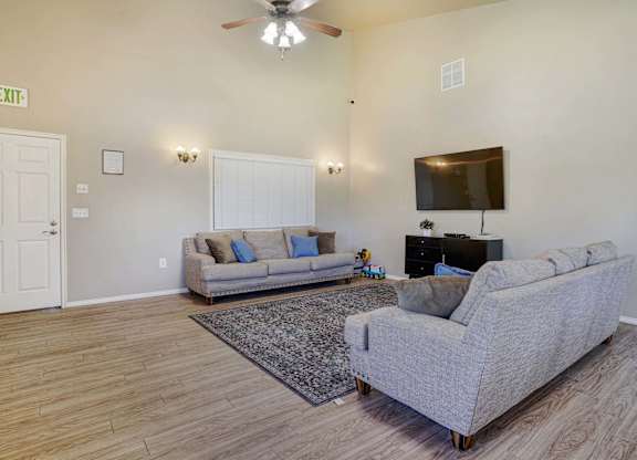 A living room with a grey couch, a rug, a ceiling fan, and a flat screen TV mounted on the wall.