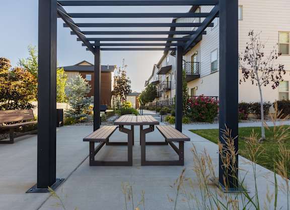 A wooden picnic table is under a black metal pergola in a courtyard.