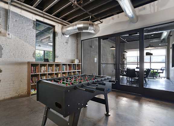 A foosball table is in the middle of a room with a brick wall and a bookshelf.