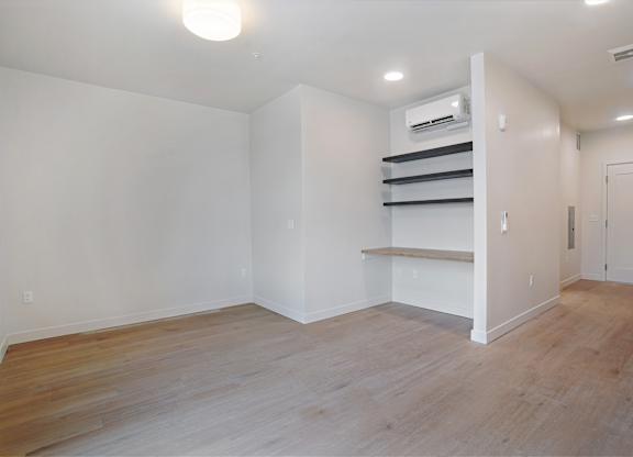 A room with a wooden floor and a white wall with a shelf and a door.