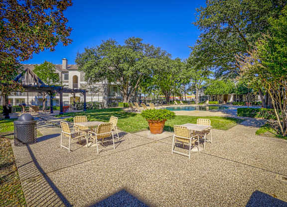 Outdoor lounge area at Saxony at Chase Oaks Apartments in Dallas, TX, featuring a shaded pergola with seating, patio tables with chairs, a lush green lawn, and a sparkling swimming pool surrounded by mature trees and apartment buildings.