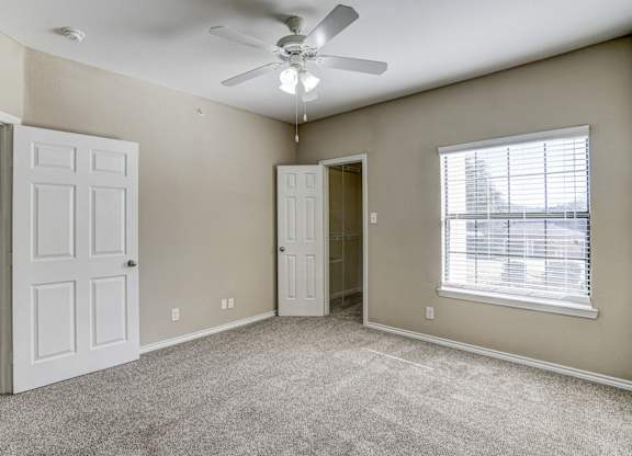 Spacious bedroom at Saxony at Chase Oaks Apartments in Dallas, TX, featuring neutral wall tones, plush carpeting, a ceiling fan with light, large window with blinds, and access to a walk-in closet through an open door.