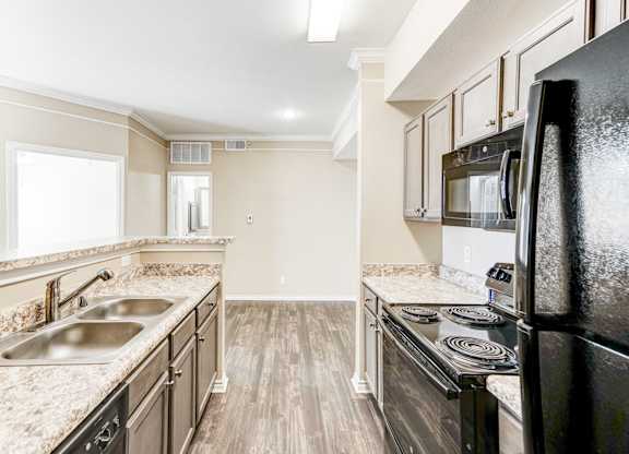 Modern kitchen at Saxony at Chase Oaks Apartments in Dallas, TX, featuring wood-style flooring, sleek black appliances, light granite countertops, dark cabinetry with brushed nickel hardware, a double stainless-steel sink, and an open layout with a view into the living area.
