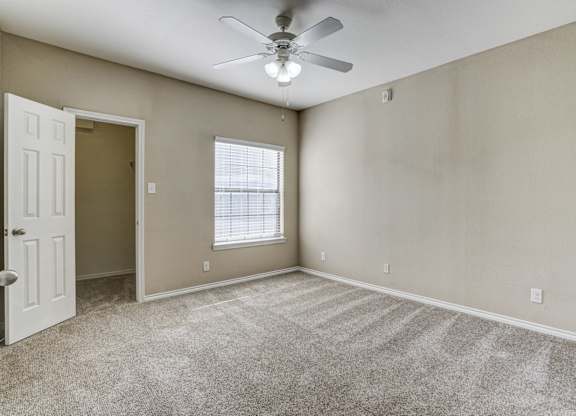 Cozy bedroom at Saxony at Chase Oaks Apartments in Dallas, TX, featuring plush carpeting, a large window with blinds letting in natural light, neutral-colored walls, a ceiling fan with light, and an open door leading to a walk-in closet.