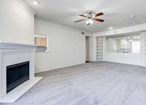 A large living room at Saxony at Chase Oaks Apartments in Dallas, TX, featuring built-in shelving, a ceiling fan, and carpeted floors, with a view of the dining and kitchen areas.
