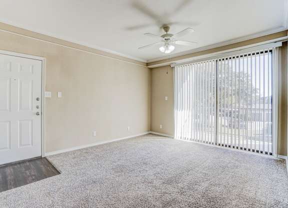 Entrance view of the living area at Saxony at Chase Oaks Apartments in Dallas, TX, showing beige walls, a white front door, and large windows with vertical blinds.