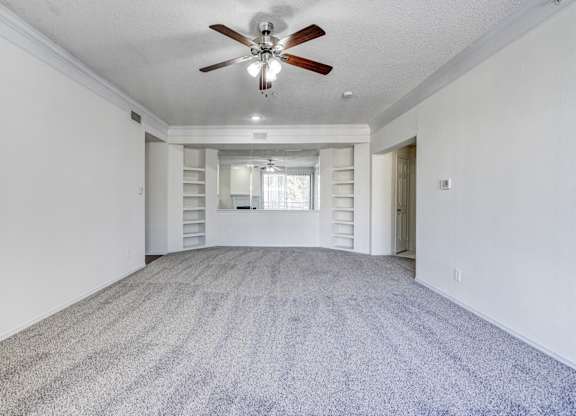 Spacious living area at Saxony at Chase Oaks Apartments in Dallas, TX, showcasing plush carpeted flooring, a ceiling fan with light, built-in shelving on either side of a mirrored accent wall, and a bright neutral color palette.