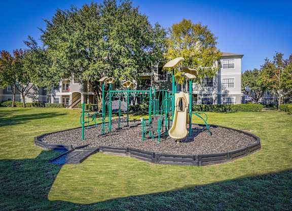 A playground surrounded by grassy lawns and mature trees at Saxony at Chase Oaks Apartments in Dallas, TX, offering a family-friendly recreational space.