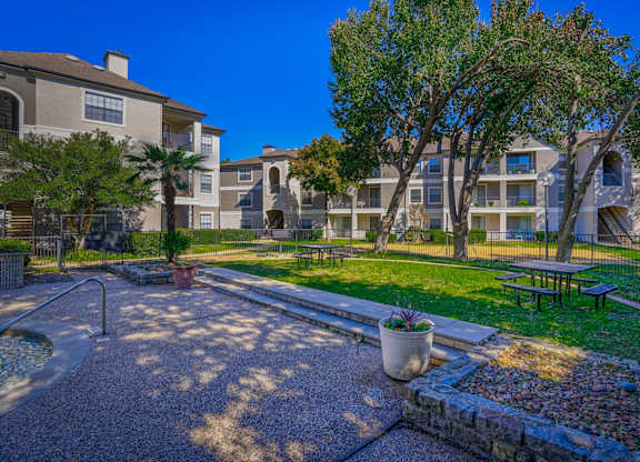 A spacious outdoor picnic area at Saxony at Chase Oaks Apartments in Dallas, TX, featuring picnic tables, a grassy lawn, and trees, offering a peaceful space for residents to gather.