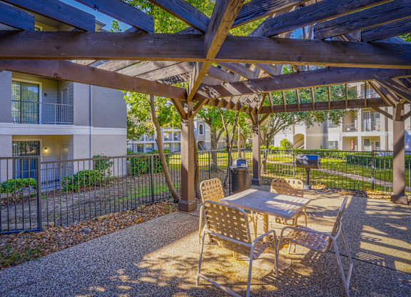 Outdoor seating area with a wooden pergola at Saxony at Chase Oaks Apartments in Dallas, TX, featuring a table and chairs, surrounded by lush landscaping and views of apartment balconies.