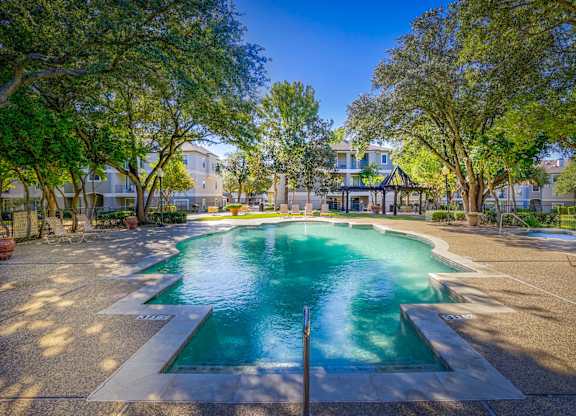 Sparkling swimming pool surrounded by a sun deck with lounge chairs and mature trees at Saxony at Chase Oaks Apartments in Dallas, TX. The area includes a shaded pergola and is framed by residential apartment buildings in the background.