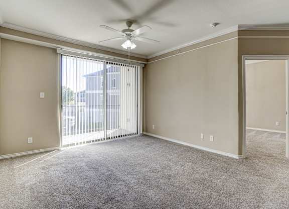 Bright and spacious living room at Saxony at Chase Oaks Apartments, featuring neutral carpeting, beige walls, crown molding, a ceiling fan, and large sliding glass doors with vertical blinds leading to a private balcony.