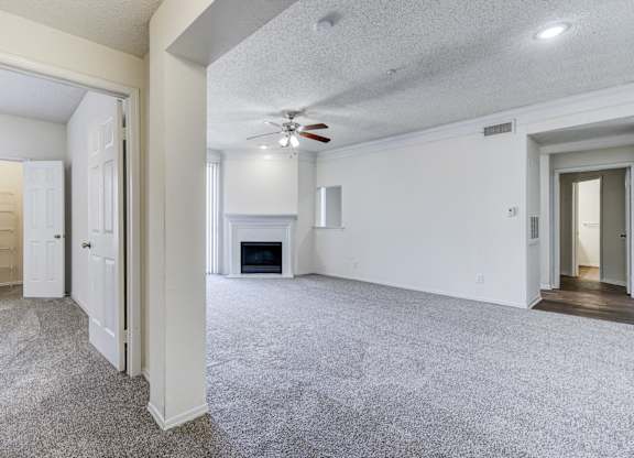 A spacious living room at Saxony at Chase Oaks Apartments in Dallas, TX, with carpeted flooring, a ceiling fan, a fireplace, and large sliding glass doors leading to a balcony.