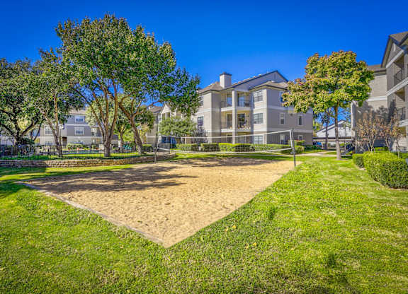 A sand volleyball court surrounded by a grassy lawn and trees at Saxony at Chase Oaks Apartments in Dallas, TX, providing an outdoor activity space for residents.