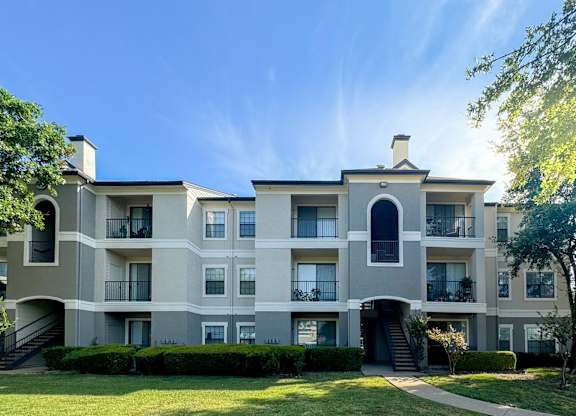 A light gray apartment building with balconies at Saxony at Chase Oaks Apartments in Dallas, TX, surrounded by manicured lawns and trees under a clear blue sky.