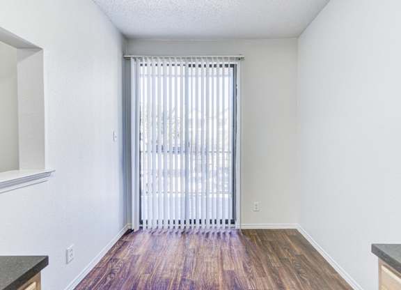 A dining area with wood-style flooring at Saxony at Chase Oaks Apartments in Dallas, TX, featuring a large sliding glass door with vertical blinds that opens to a balcony.