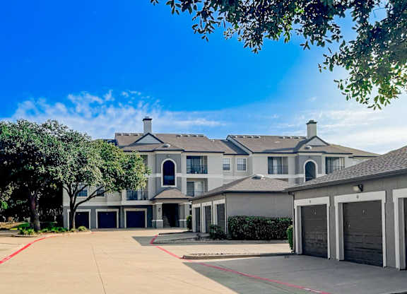 Exterior view of Saxony at Chase Oaks Apartments in Dallas, TX, showcasing three-story buildings with arched entryways, private balconies, and attached garages, surrounded by mature trees and a spacious driveway.