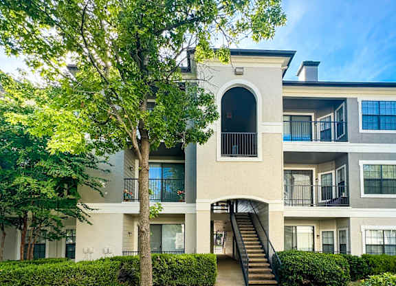 A three-story apartment building at Saxony at Chase Oaks Apartments in Dallas, TX, surrounded by a green lawn, clear blue skies, and neatly trimmed shrubs.