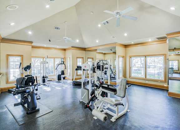 Fitness center at Saxony at Chase Oaks Apartments in Dallas, TX, featuring modern exercise equipment, a mirrored wall, vaulted ceilings with ceiling fans, and large windows letting in natural light.