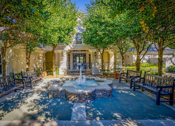 A tranquil courtyard at Saxony at Chase Oaks Apartments in Dallas, TX, featuring a central fountain surrounded by benches, lush green trees, and a shaded pathway leading to the entrance of the clubhouse.