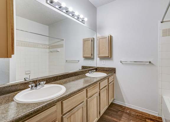 Bright and spacious bathroom at Saxony at Chase Oaks Apartments in Dallas, TX, featuring dual sinks, a large mirror, and wood-style flooring.