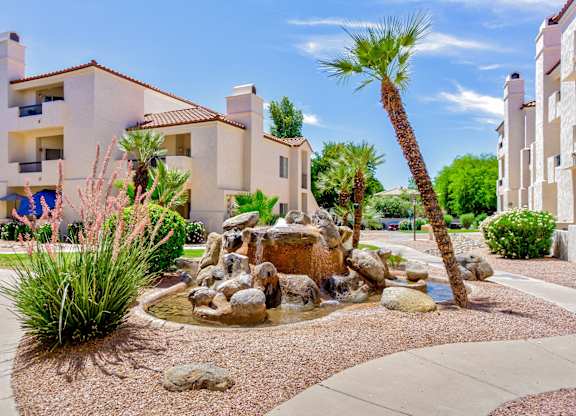 A fountain in the middle of a garden with a palm tree and apartment buildings in the background.