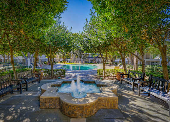 A shaded courtyard at Saxony at Chase Oaks Apartments in Dallas, TX, featuring a decorative fountain, benches, and lush greenery, with views of the pool area in the distance.