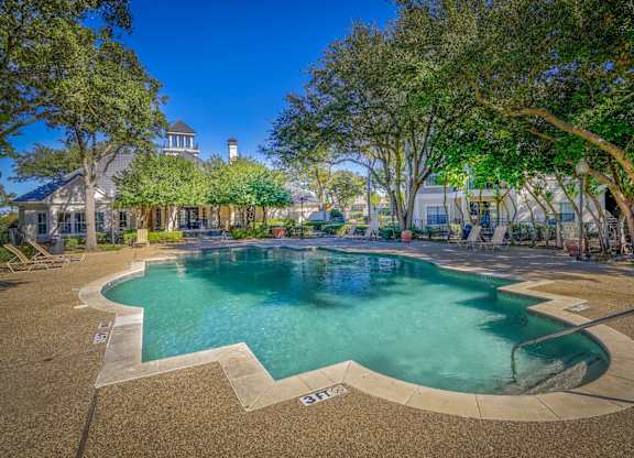 Outdoor pool area at Saxony at Chase Oaks Apartments in Dallas, TX, featuring a clear turquoise swimming pool, a sun deck with lounge chairs, mature trees for shade, and a charming clubhouse in the background with a turret-style roof.