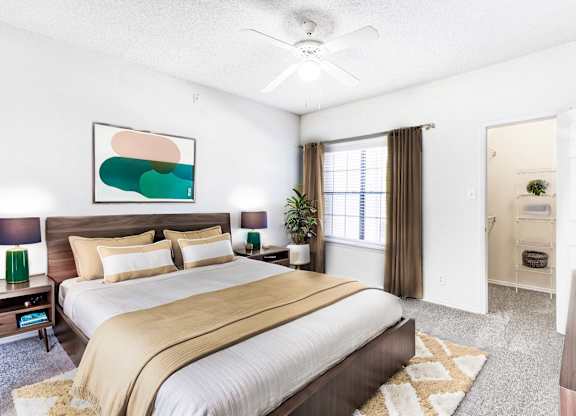 A staged bedroom at Saxony at Chase Oaks Apartments in Dallas, TX, showcasing a modern bed with stylish bedding, side tables, a ceiling fan, and a walk-in closet with shelving.