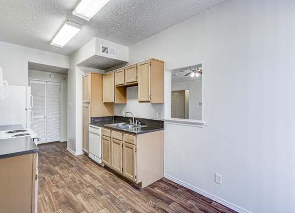 A well-lit kitchen at Saxony at Chase Oaks Apartments in Dallas, TX, featuring wooden cabinets, black countertops, a white dishwasher, a stove, and a refrigerator, with wood-style flooring and a view of a hallway.