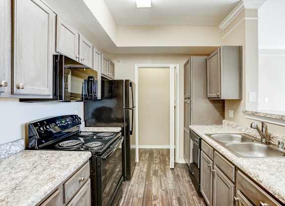 Contemporary kitchen at Saxony at Chase Oaks Apartments in Dallas, TX, featuring sleek dark cabinetry, granite-style countertops, stainless steel double sinks, and modern black appliances, including a stove, microwave, and refrigerator.