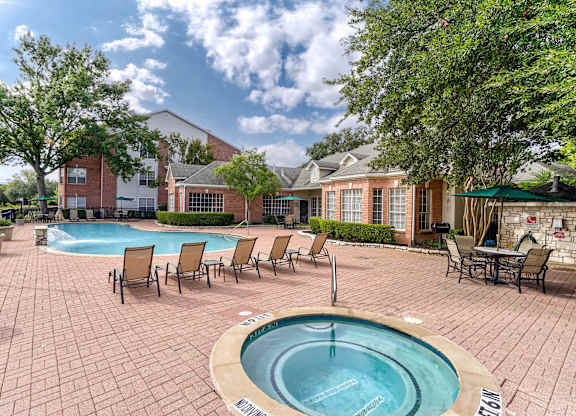 A pool with a hot tub in the middle of a brick patio surrounded by chairs.