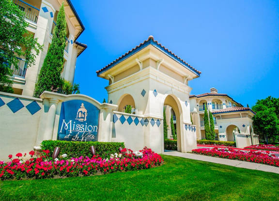 Community entrance with florals and luscious grass at Mission at La Villita Apartments in Irving, TX offers 1, 2 & 3 bedroom apartment homes with appliances.