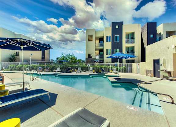 Pool with buildings in the background and patio umbrella up front. Four lounge chairs line the pool.