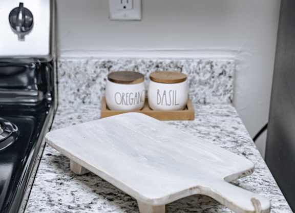 A white cutting board with two jars of organic and basil on a marble countertop.