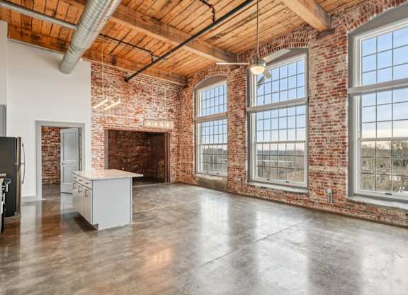 Unstaged living room in Converse Mill Lofts in Spartanburg, SC with high ceilings, exposed beams, brick walls, large windows and a kitchen with stainless steel appliances.