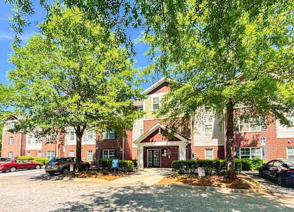 A building with a red brick facade and a large front yard with trees and a gravel area.