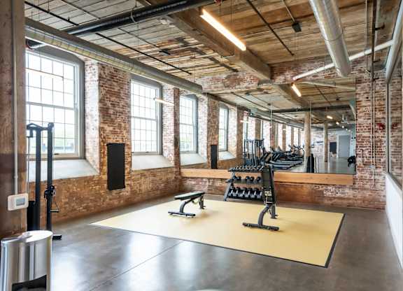 Loft Apartments for Rent in Spartanburg, SC - Converse Mill Lofts - Fitness Center with High Ceiling, Exposed Ductwork and Brick Walls, Large Windows, Large Mirror, Free Weights, and Cardio Equipment.