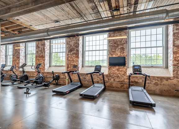 Loft Apartments for Rent in Spartanburg, SC - Converse Mill Lofts - Fitness Center with High Ceiling, Exposed Ductwork and Brick Walls, Large Windows, Large Mirror, Free Weights, and Cardio Equipment.