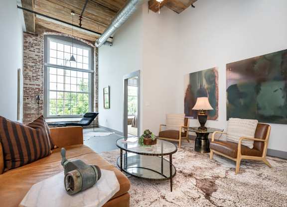 Loft Apartments Near Downtown Spartanburg, SC - Converse Mill Lofts - Living Room with High Ceiling, Large Window, Chaise Lounge, Pendant Lighting, Couch, Coffee Table, Armchairs, and Paintings.