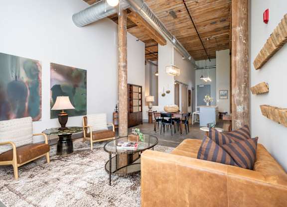 Loft Apartments Near Downtown Spartanburg, SC - Converse Mill Lofts - Living Room with High Ceiling, Large Window, Chaise Lounge, Pendant Lighting, Couch, Coffee Table, Armchairs, and Paintings.